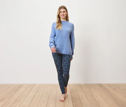 Eine Frau geht barfuß auf einem Holzboden und trägt einen blauen Pullover und gemusterte Leggings.