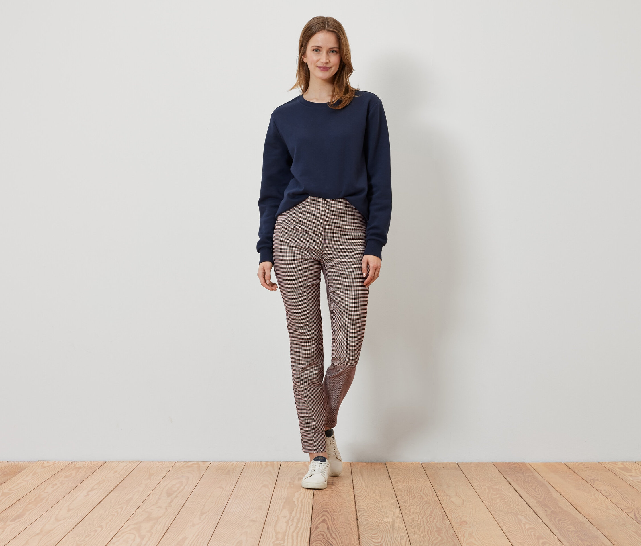 Eine Frau steht auf einem Holzboden und trägt ein dunkelblaues Sweatshirt und eine braun karierte Hose.
