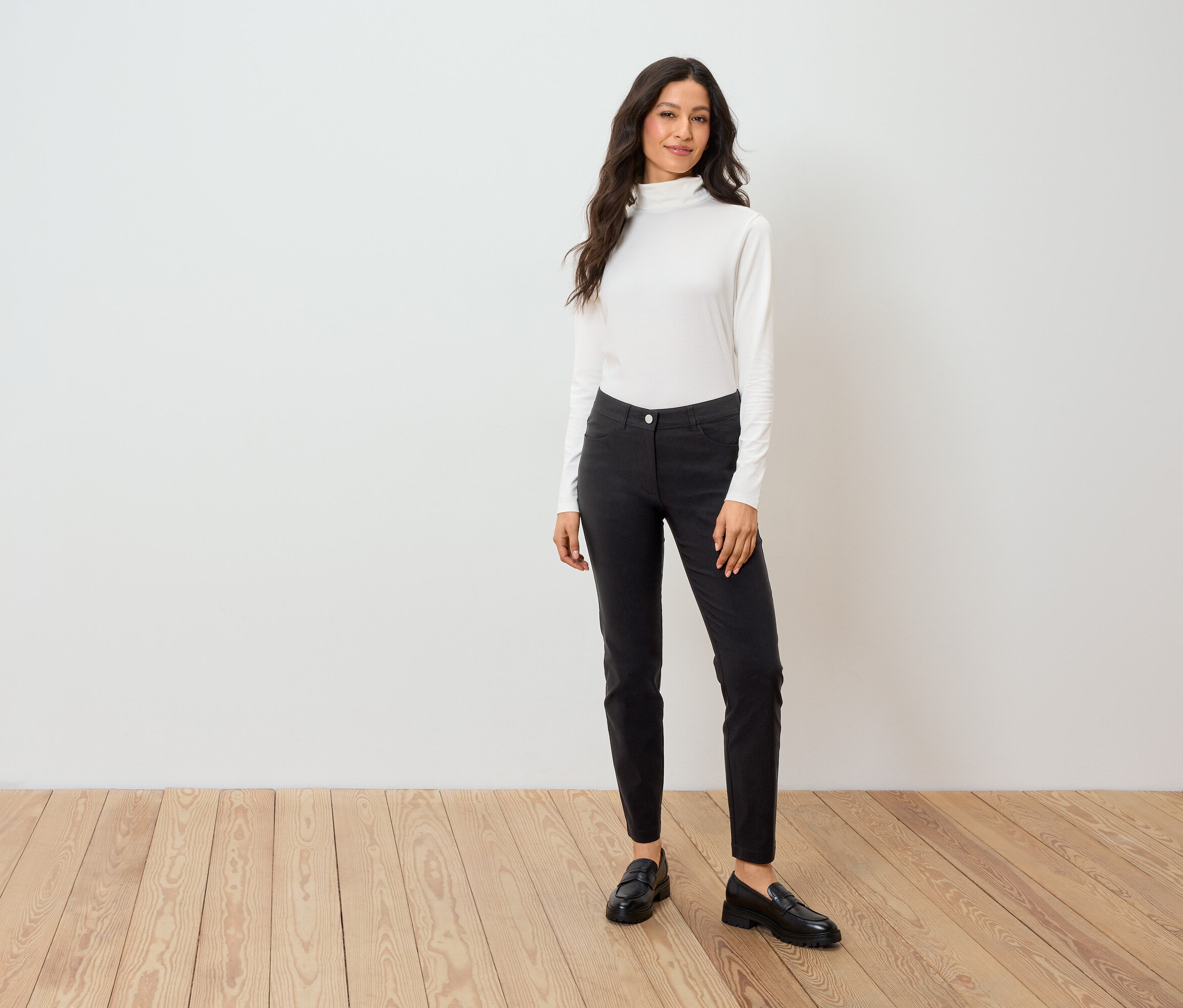 Frau steht auf Holzboden und trägt einen weißen Rollkragenpullover, schwarze Hosen und schwarze Slipper.