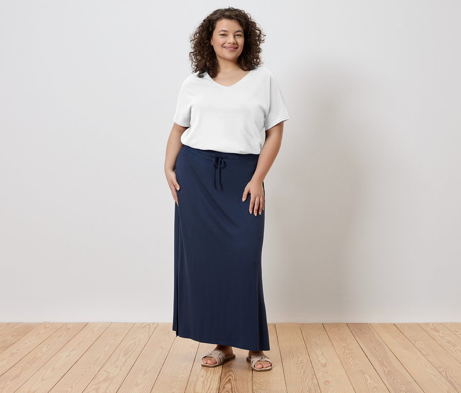 Frau steht und trägt ein weißes T-Shirt, einen langen, marineblauen Rock und Sandalen.