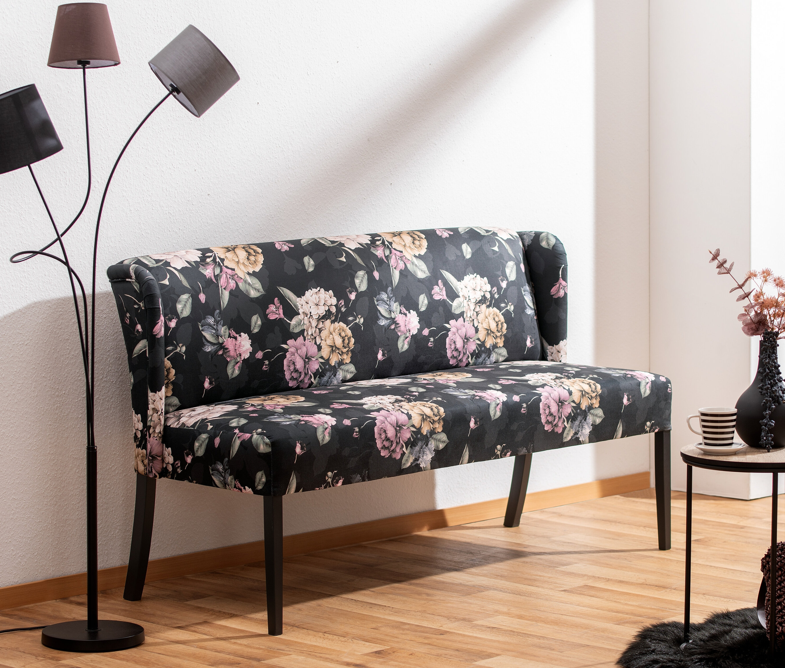 Gepolsterte »Hendrick« Bank mit Blumenmuster in Anthrazit steht neben einer Stehlampe.