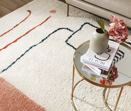Ein beiger Teppich mit roten und blauen Linien liegt unter einem goldenen Beistelltisch mit Büchern, einer Vase und rosa Blumen.