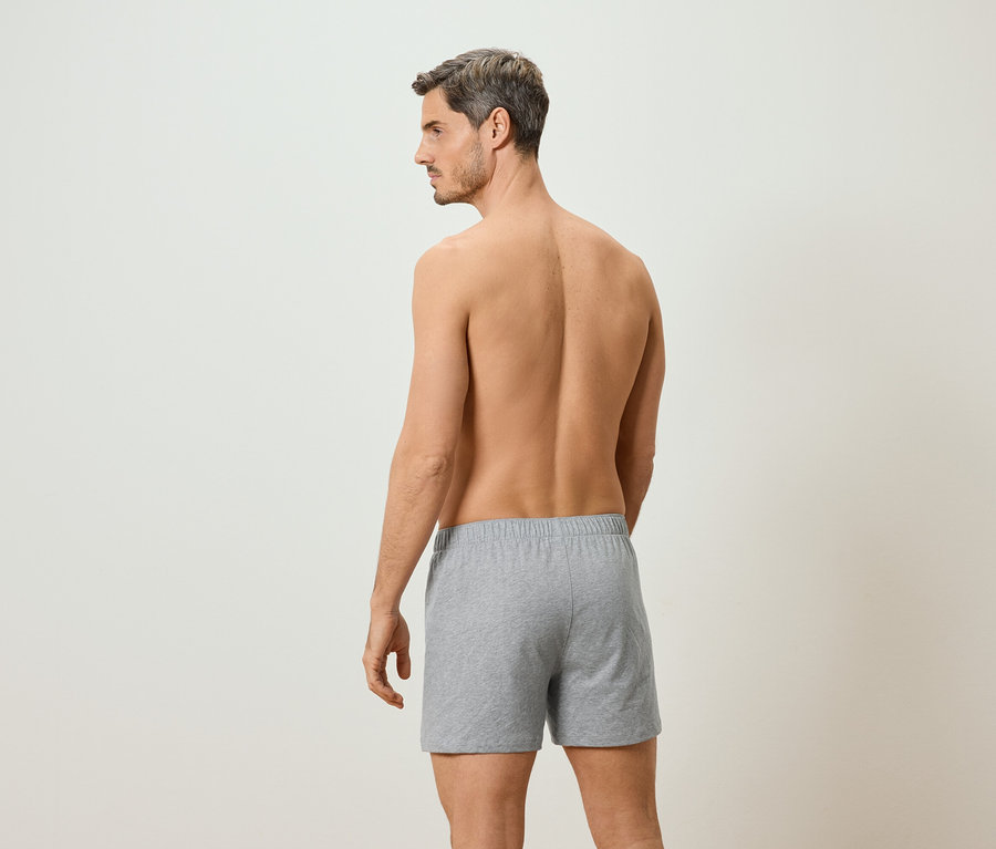 Mann mit freiem Oberkörper, von hinten zu sehen, trägt graue Boxershorts vor einem neutralen Hintergrund.