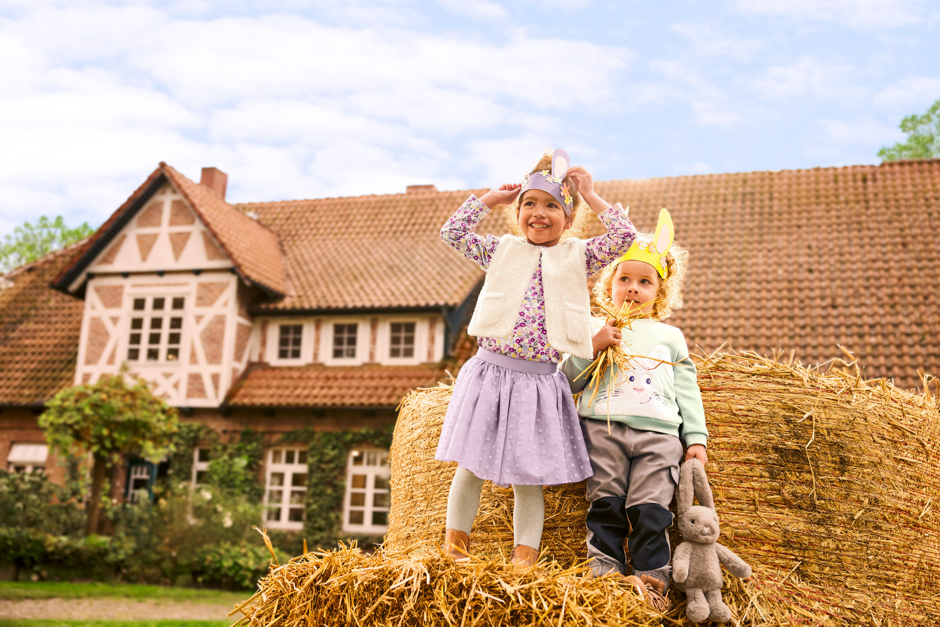 Zwei Mädchen stehen auf einem Strohballen vor einem Haus. Das Mädchen links trägt Lederstiefel, eine Kinder-Teddyweste, ein mintfarbenes Sweatshirt mit angerauter Innenseite, einen Kinder-Tüllrock, 5 Kinder-Strumpfhosen und 2 Kinder-Langarmshirts mit Blumenapplikation. Das Mädchen rechts hält ein Plüschtier »Hase« und trägt eine braune Kinder-Outdoorhose.