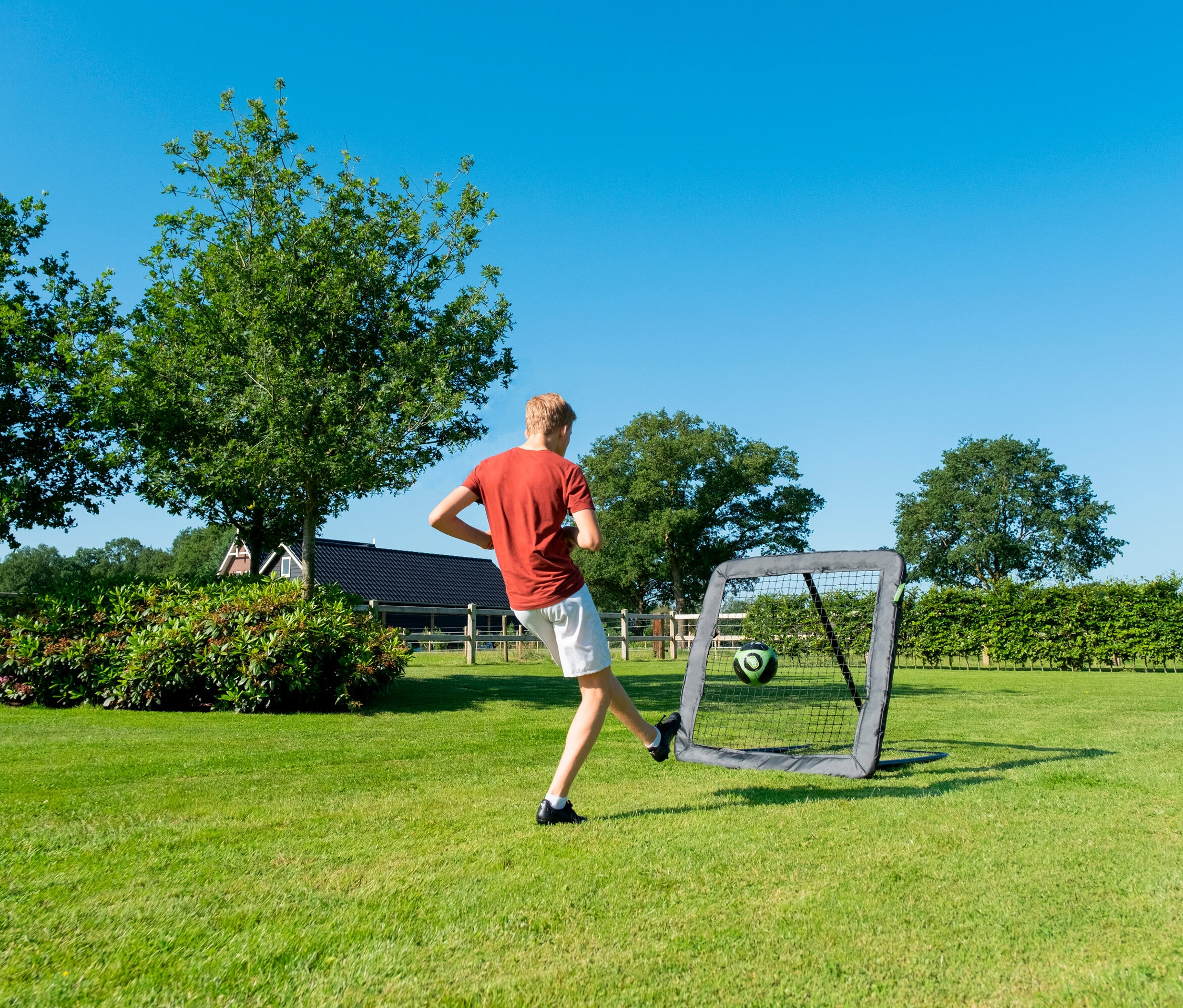 Auf einer Wiese schießt ein Mann in ein graues Fußballtor, im Hintergrund Bäume und ein Haus.