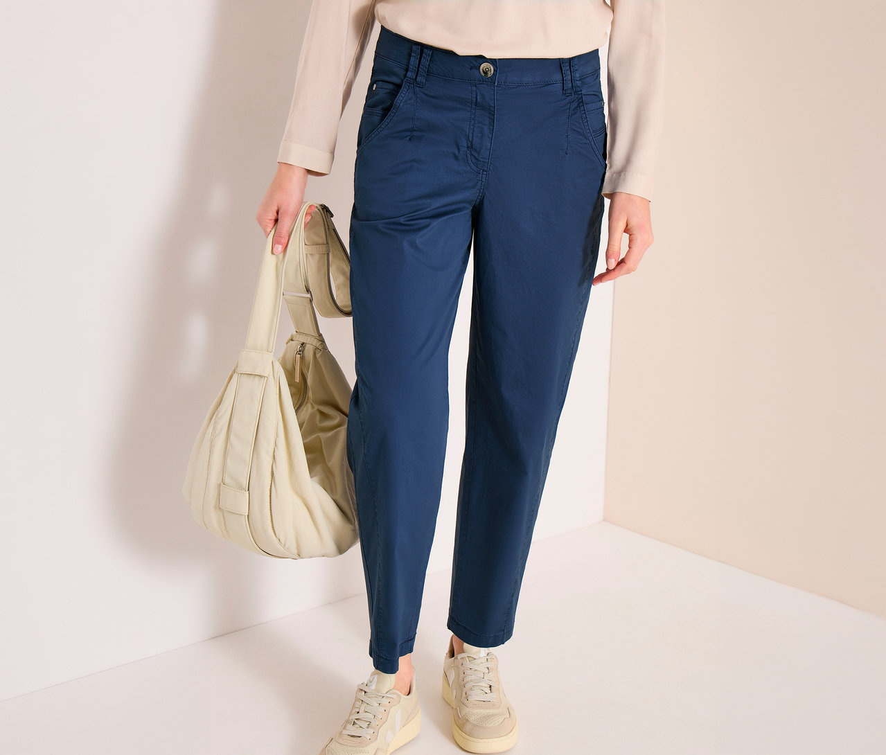 Eine Person trägt blaue CECIL Barrel »Scarlett« Hose, ein helles Oberteil und helle Sneaker, während sie eine gepolsterte, helle Umhängetasche hält.