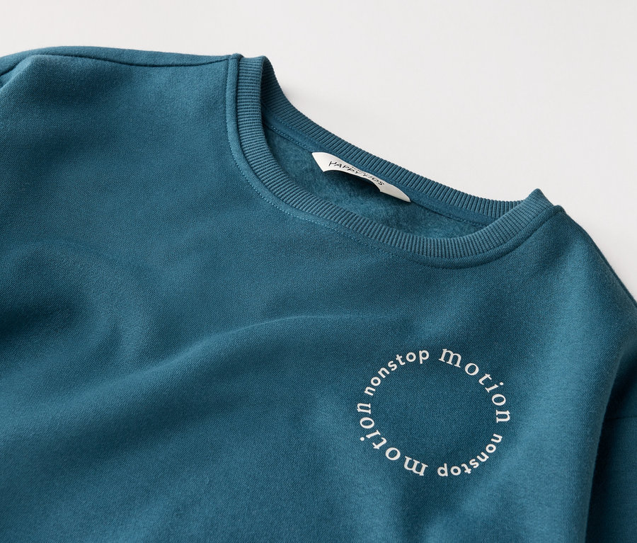 Nahaufnahme eines petrolfarbenen Sweatshirts mit der Aufschrift "nonstop motion" in einem Kreis.