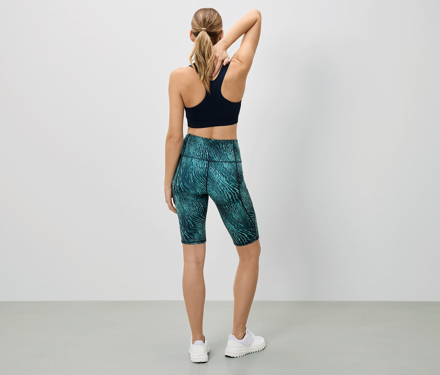 Eine Frau steht mit dem Rücken zur Kamera, trägt einen schwarzen Sport-BH und kurze, gemusterte Leggings.