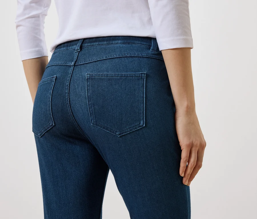 Rückansicht einer Frau mit weißem Oberteil und blauen Jeans mit Gesäßtaschen vor einem weißen Hintergrund.