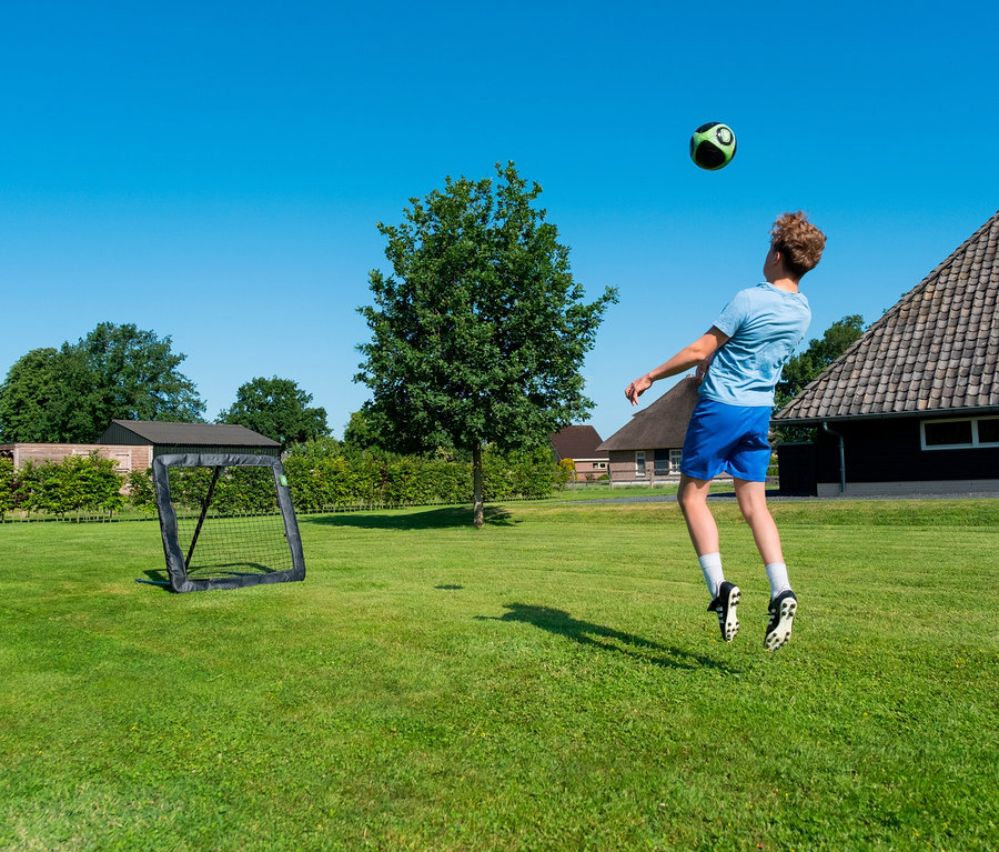 Ein Jugendlicher köpft einen Fußball auf einer grünen Wiese vor einem Fußballtor und Häusern unter blauem Himmel.