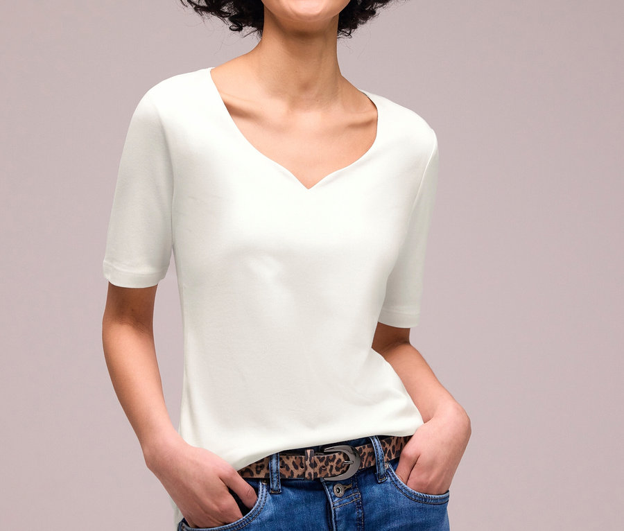 Lachende Frau mit lockigem Haar trägt ein weißes Street One Kurzarmshirt, blaue Jeans und einen Gürtel mit Leopardenmuster, die Hände in den Taschen.