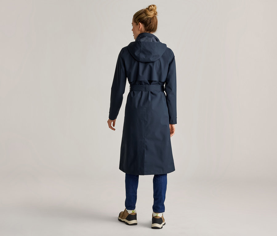 Rückansicht einer Frau mit Duttfrisur, die einen langen, marineblauen Trenchcoat mit Kapuze, blaue Jeans und braune Schuhe trägt.