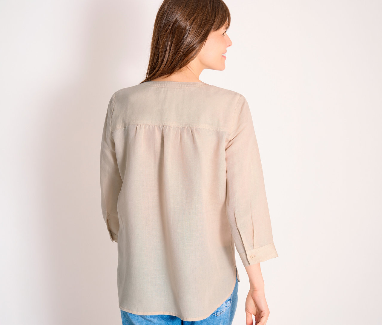 Rückansicht einer Frau mit langen braunen Haaren, die eine hellbeige, langärmlige Bluse und blaue Jeans trägt.