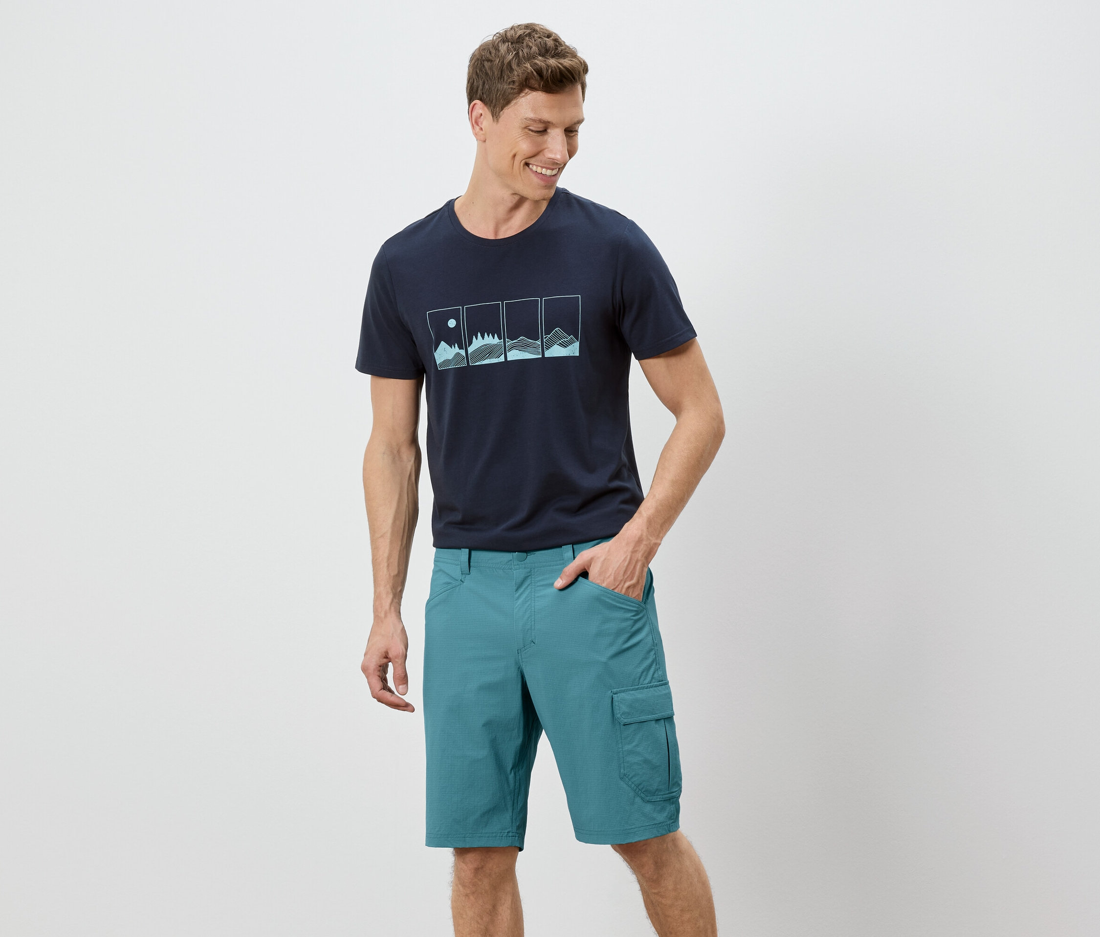 Ein Mann steht mit einer Hand in der Tasche vor einem weißen Hintergrund. Er trägt ein marineblaues T-Shirt mit einer Grafik und grüne Shorts.