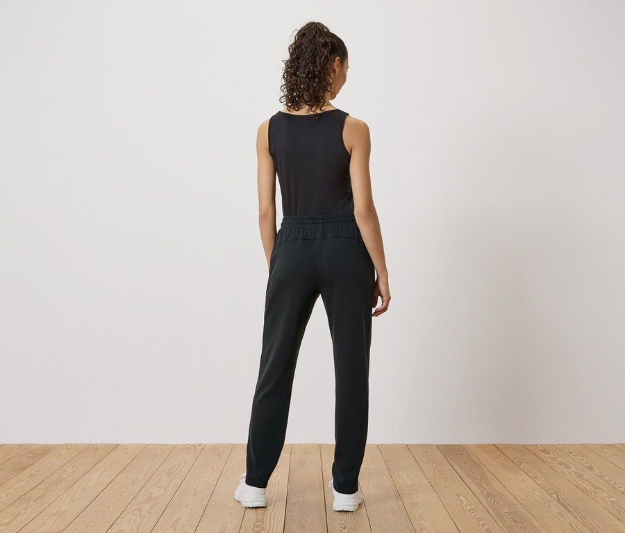 Rückansicht einer Frau mit lockigem Haar, die ein schwarzes Tanktop, eine schwarze Hose und weiße Turnschuhe trägt.