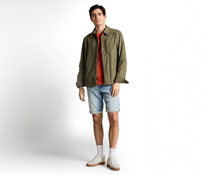 Mann in olivgrüner Jacke und Tom Tailor TTJOSH REGULAR SLIM Jeansshorts in Light Stone Blue Denim, dazu T-Shirt, Socken und Sneaker.
