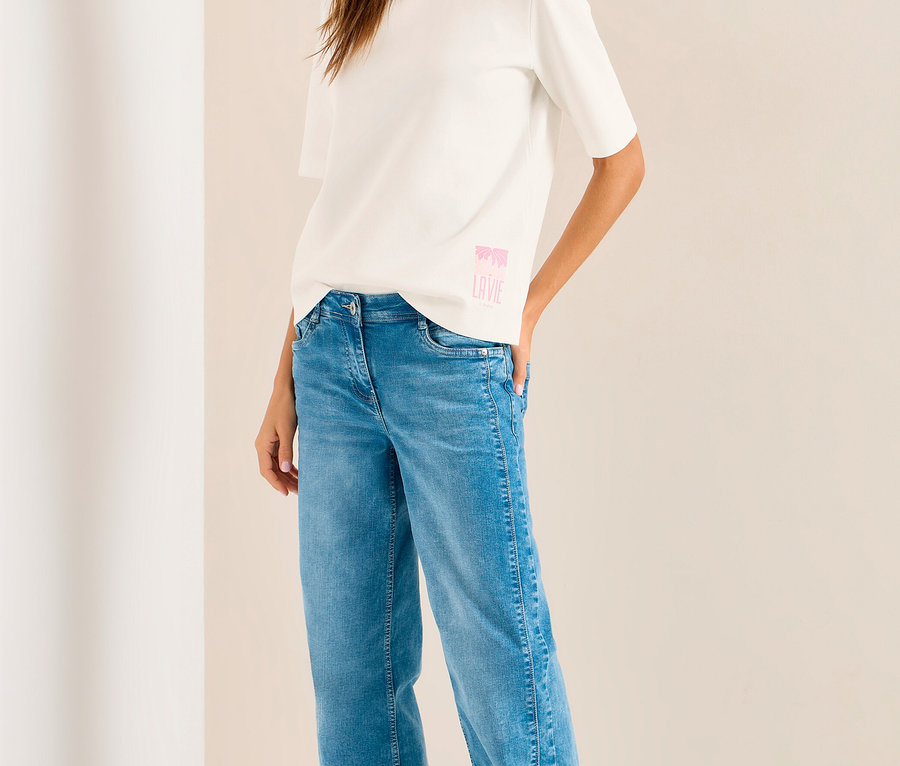 Lächelnde Frau in weißem CECIL Sweatshirt mit pinkem Aufdruck, blauer Jeans, weißen Sneakern und hellvioletter Kappe.