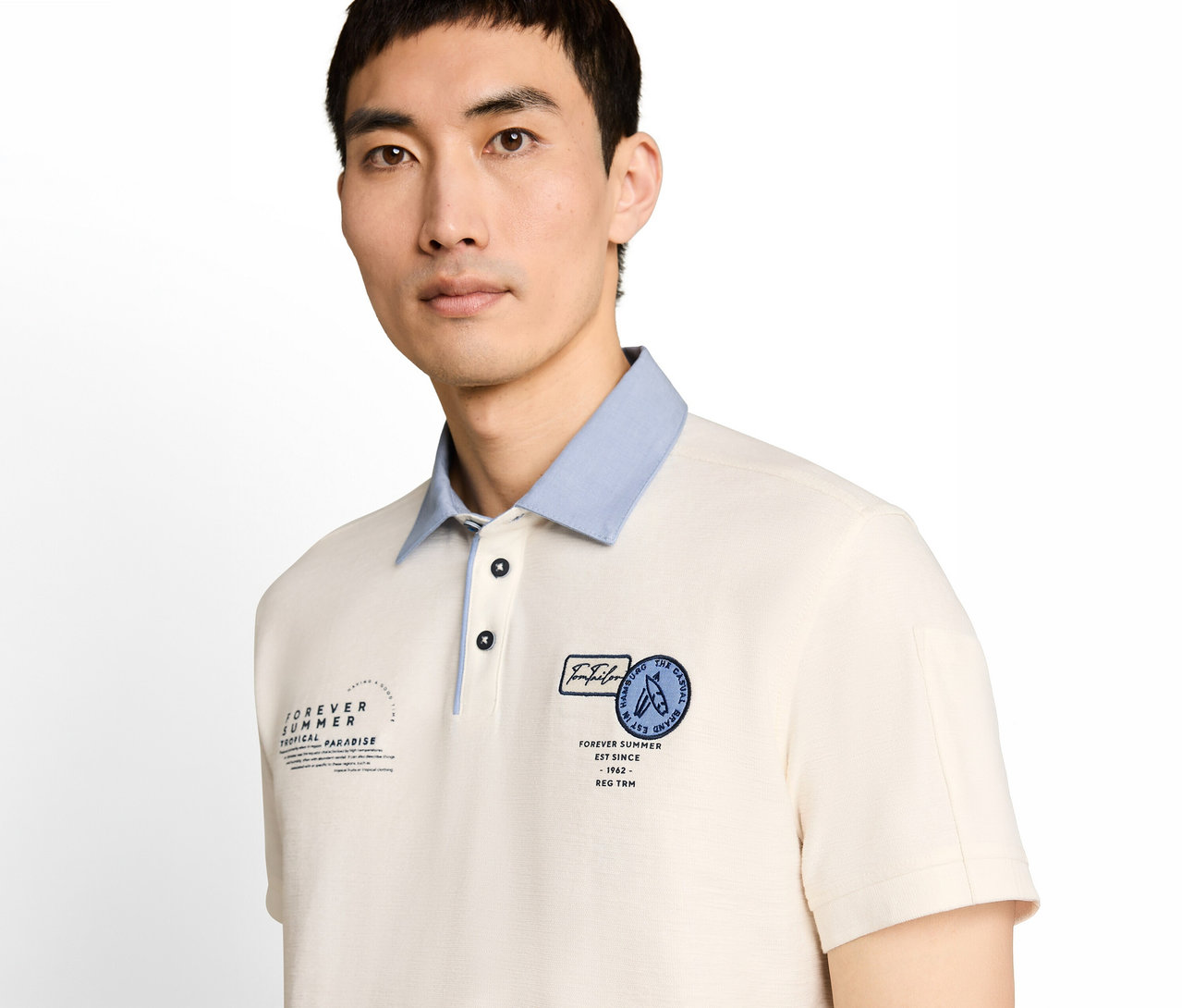 Ein Mann trägt ein cremefarbenes Poloshirt mit hellblauem Kragen und zwei dunklen Knöpfen. Das Shirt zeigt links den Schriftzug „FOREVER SUMMER TROPICAL PARADISE“ und rechts einen runden „TOM TAILOR“-Aufnäher mit Surfbrettern.