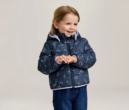 Lächelndes Mädchen mit blonder Frisur trägt eine marineblaue, gesteppte Jacke mit Kapuze und Sternenmuster und blaue Jeans.