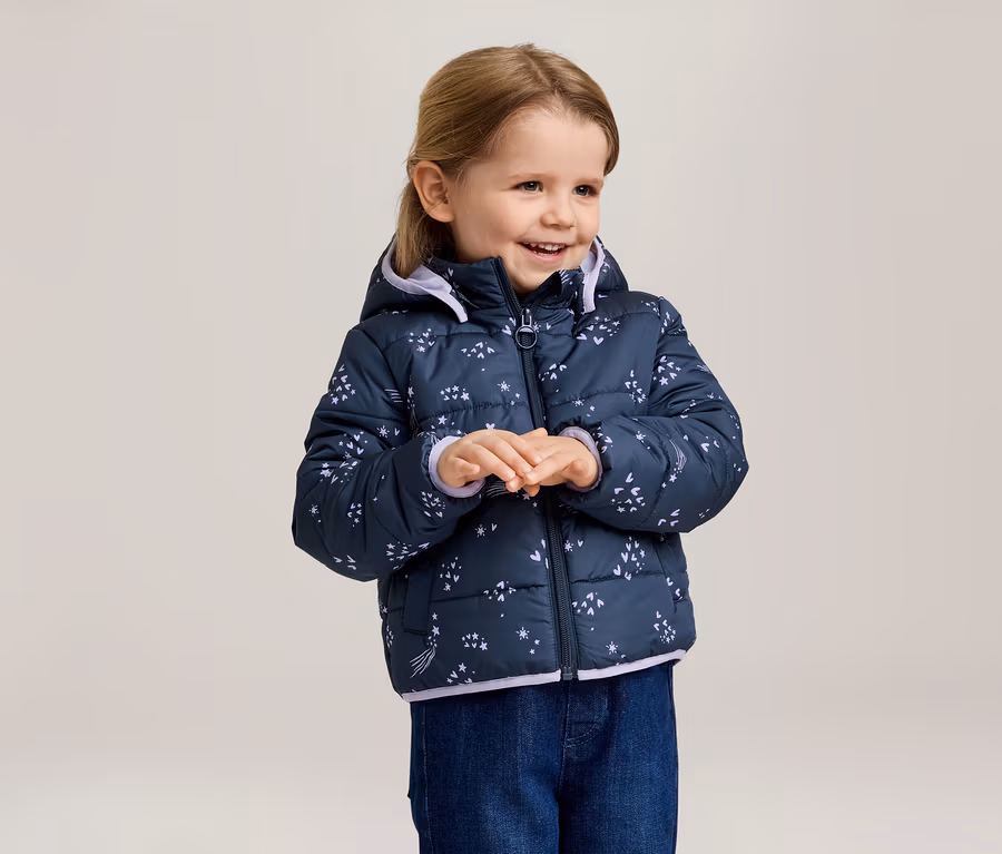 Lächelndes Mädchen mit blonder Frisur trägt eine marineblaue, gesteppte Jacke mit Kapuze und Sternenmuster und blaue Jeans.