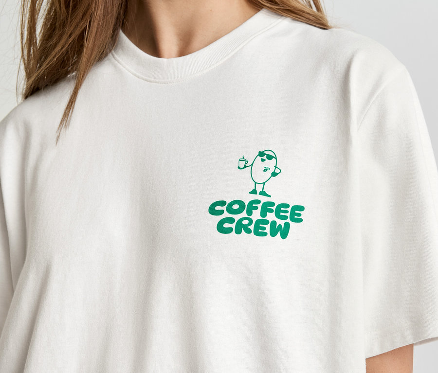 Nahaufnahme einer Person, die ein helles T-Shirt mit dem grünen Aufdruck "Coffee Crew" und einer Cartoon-Kaffeebohne trägt.