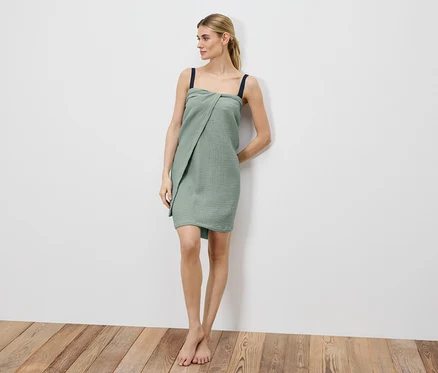 Frau steht barfuß an einer weißen Wand, gehüllt in ein grünes Musselin-Tuchkleid mit dunklen Trägern.