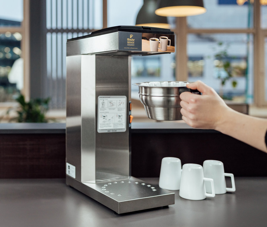 Eine Hand hält einen Edelstahlfilter gefüllt mit weißen Papierfiltern über eine Kaffeemaschine. Im Hintergrund stehen drei weiße Tassen. Im Fokus: Profi-Filterkaffeemaschine »Tchibo Businessline«, ohne Kanne