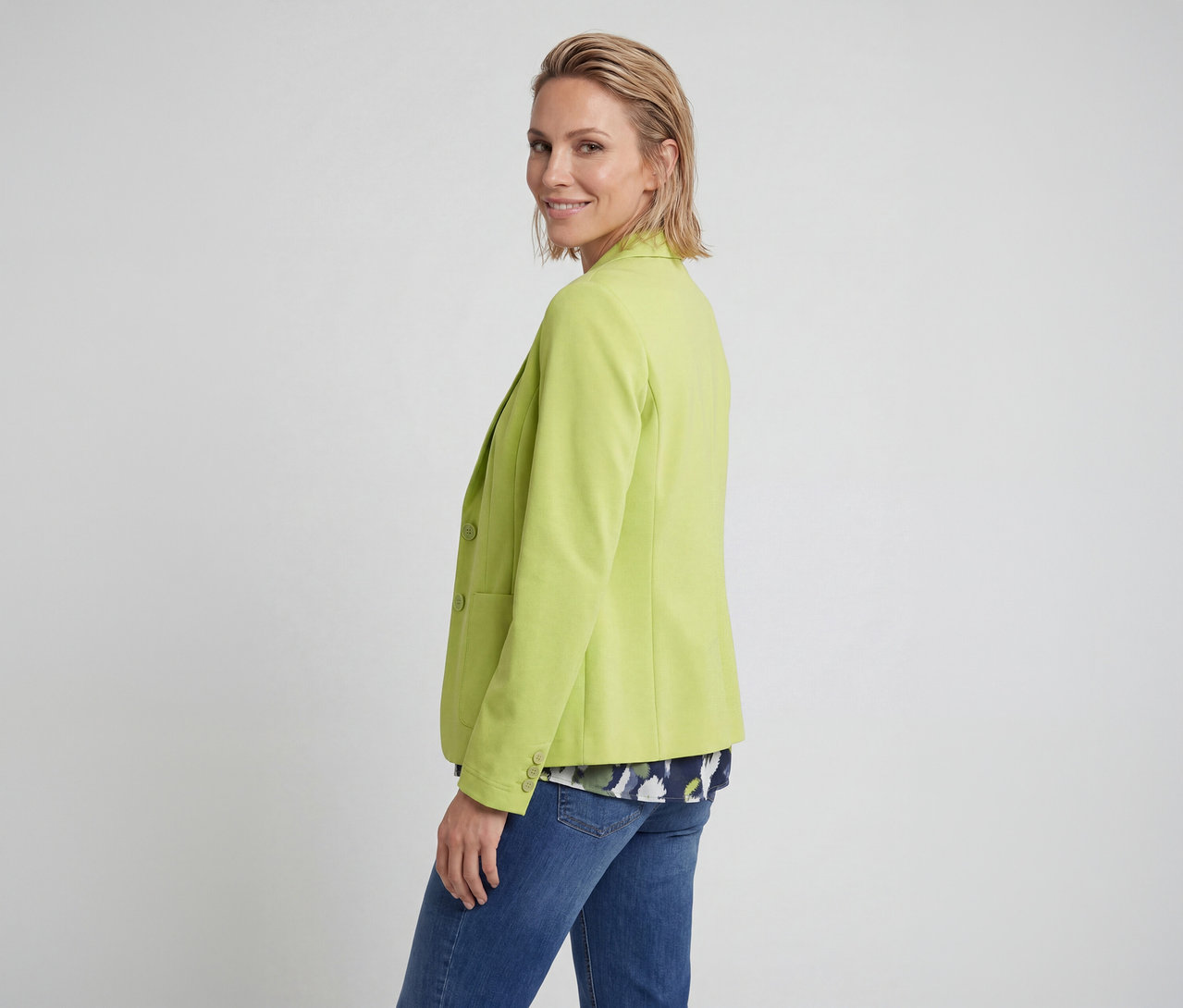 Frau in einem limettengrünen Blazer, einer gemusterten Bluse und blauen Jeans, die sich nach rechts dreht und in die Kamera lächelt.