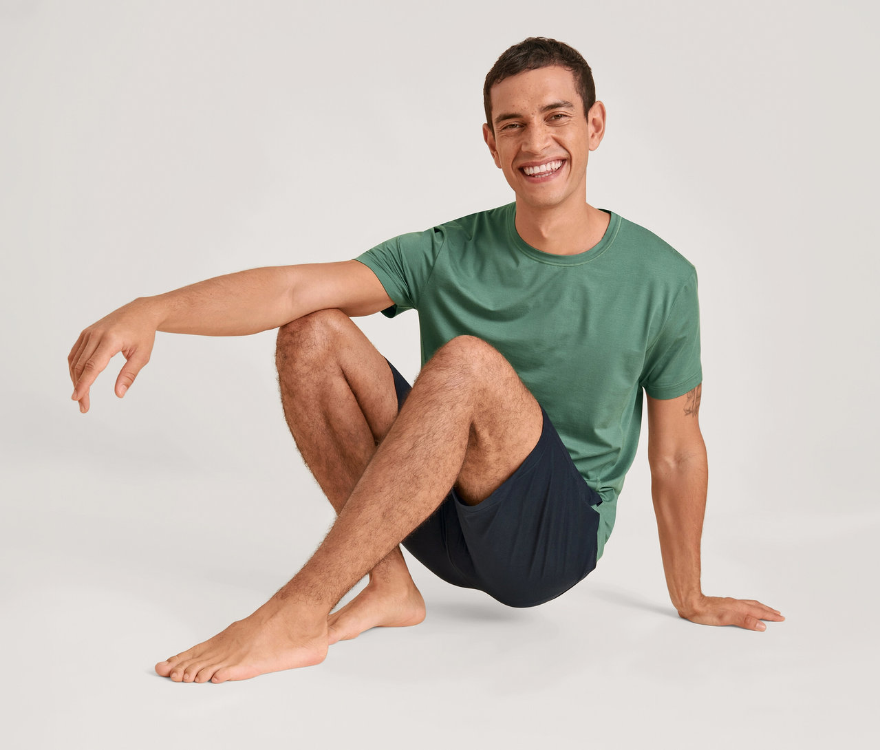 Ein Mann mit kurzem dunklem Haar, der ein grünes T-Shirt und dunkle Shorts trägt, sitzt barfuß auf einem hellen Boden und lächelt in die Kamera. Sein linker Arm ruht auf seinem angewinkelten linken Bein, seine rechte Hand liegt auf dem Boden.