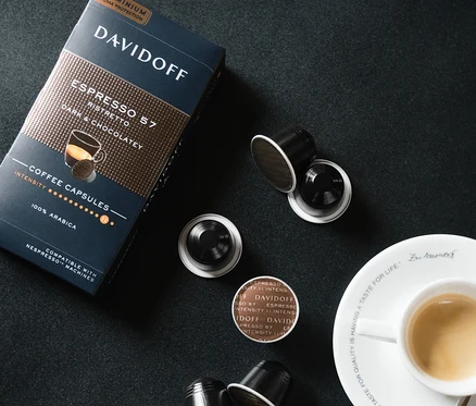 Eine Packung und mehrere Kapseln DAVIDOFF CAFÉ Espresso 57 Ristretto neben einer Tasse Espresso auf schwarzem Hintergrund.