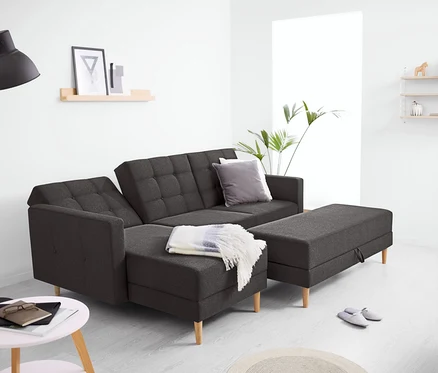 Dunkelgraues Federkern-Eckschlafsofa »Just Relax« mit Stauraumbank in einem modernen Wohnzimmer. Auf dem Sofa liegt eine helle Decke, daneben die passende Stauraumbank. Eine schwarze Stehlampe steht links, ein Beistelltisch mit Notizbuch und Brille davor. Im Hintergrund sind Regale und eine Pflanze zu sehen.