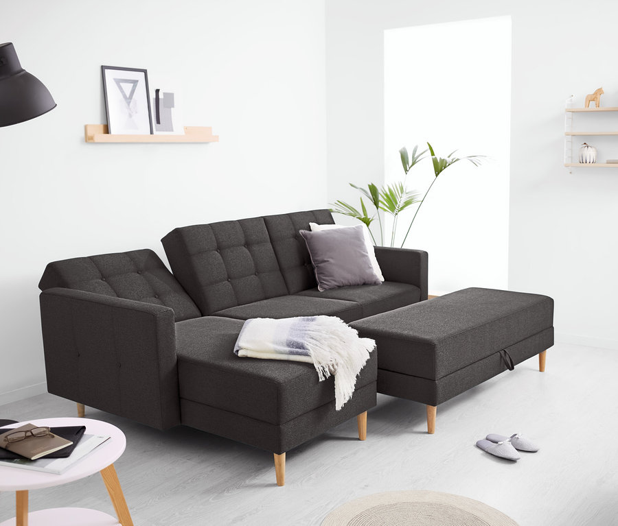 Dunkelgraues Federkern-Eckschlafsofa »Just Relax« mit Stauraumbank in einem modernen Wohnzimmer. Auf dem Sofa liegt eine helle Decke, daneben die passende Stauraumbank. Eine schwarze Stehlampe steht links, ein Beistelltisch mit Notizbuch und Brille davor. Im Hintergrund sind Regale und eine Pflanze zu sehen.