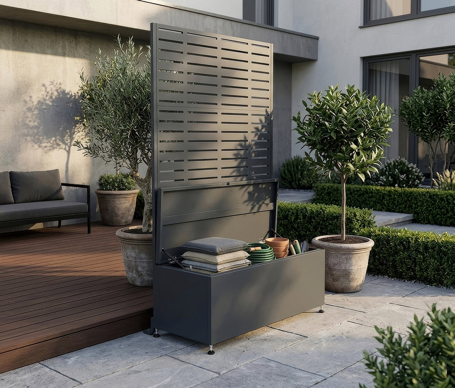Dunkelgraue Outdoor-Aufbewahrungsbox mit integriertem Lamellen-Sichtschutz, geöffnet mit gestapelten Kissen und Gartengeräten darin. Die Box steht auf einer Steinterrasse neben einer Holzterrasse, mit Topfbäumen und Hecken im Hintergrund.