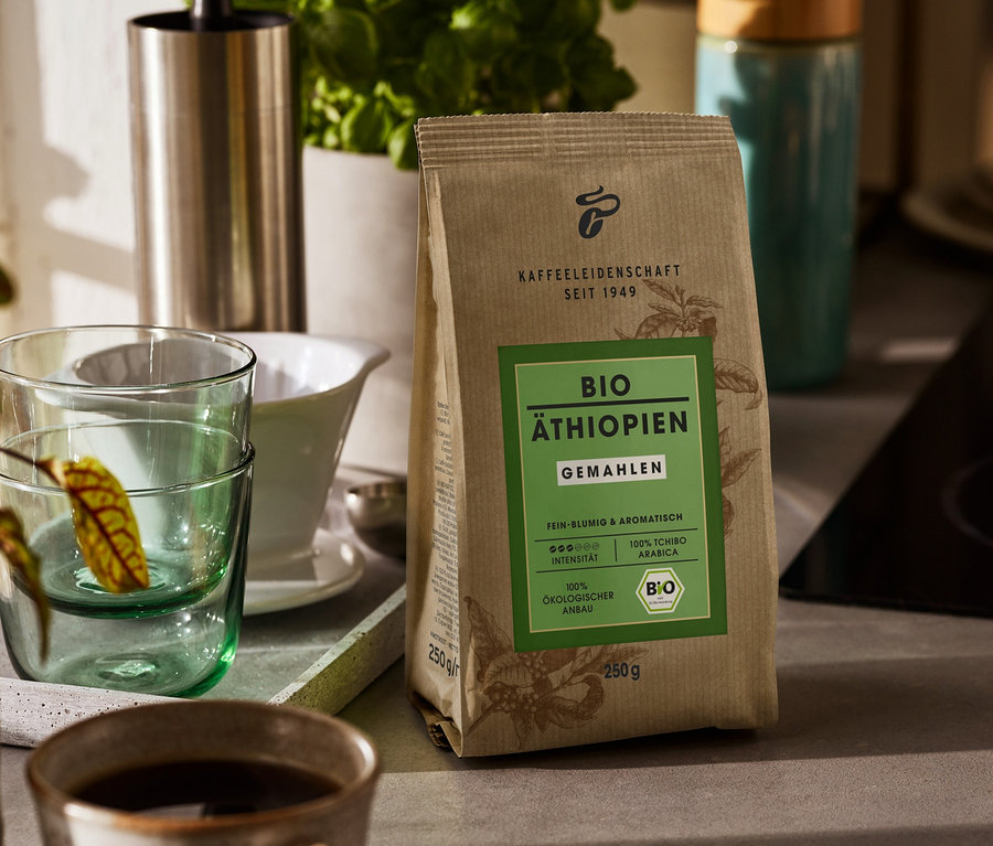 Eine Packung Bio Äthiopien - 250 g Gemahlen steht auf einer Küchentheke. Im Hintergrund befinden sich eine Kaffeemühle, ein Wasserkocher und eine Tasse Kaffee.