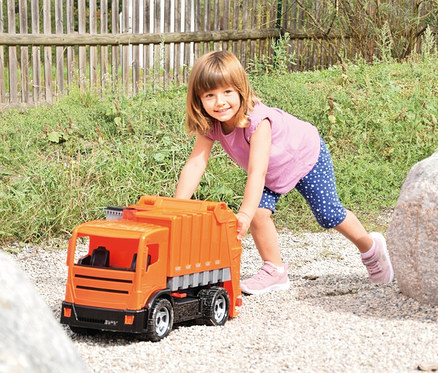 Kleines Mädchen schiebt einen orangefarbenen GIGA TRUCKS Müllwagen auf einem Kiesweg und lächelt in die Kamera. Sie trägt ein pinkes T-Shirt und blaue Leggings mit weißen Punkten. Im Hintergrund sind grüne Vegetation und ein Holzzaun zu sehen.