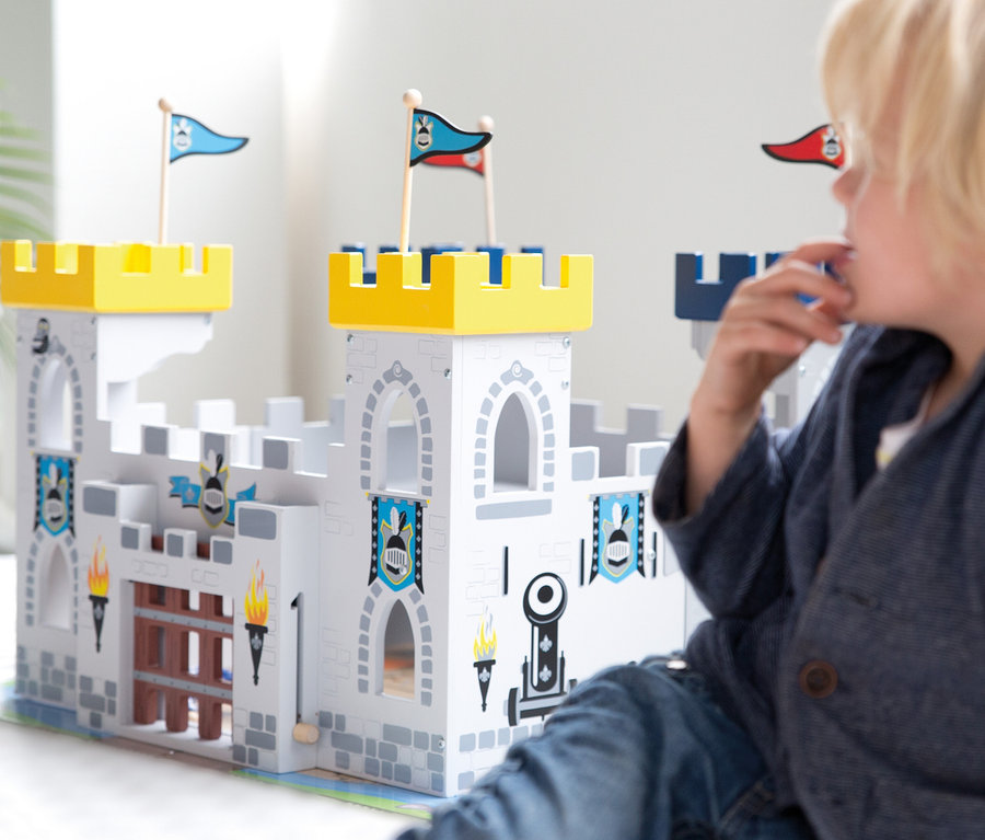 Blondes Kind in blauer Jacke sitzt vor einem weißen Holzspielzeugschloss mit gelben Zinnen, Fahnen und Zugbrücke.