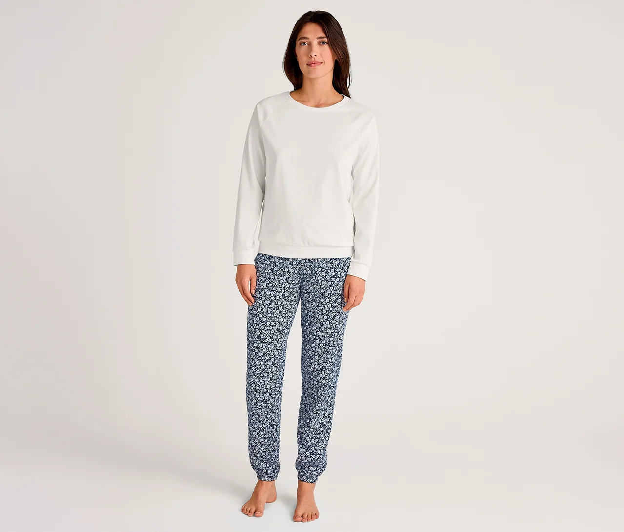 Frau steht barfuß, trägt ein weißes Langarmshirt und eine dunkelblaue Pyjamahose mit hellem Blumenmuster.