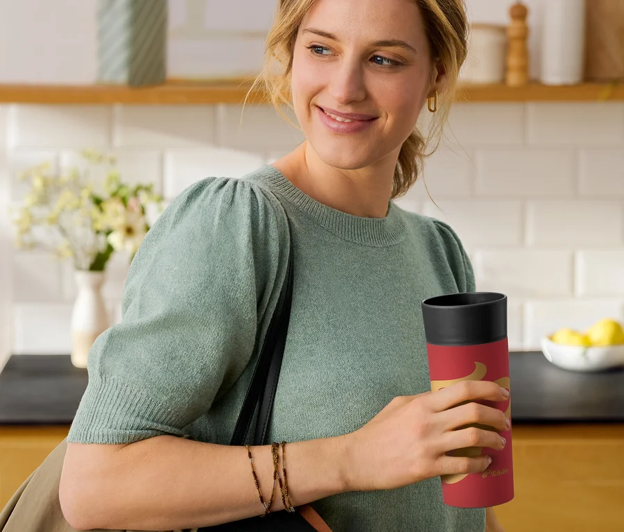 Lächelnde Frau in grünem Pullover hält einen roten Thermo Becher mit Bohnen-Design in einer Küche.