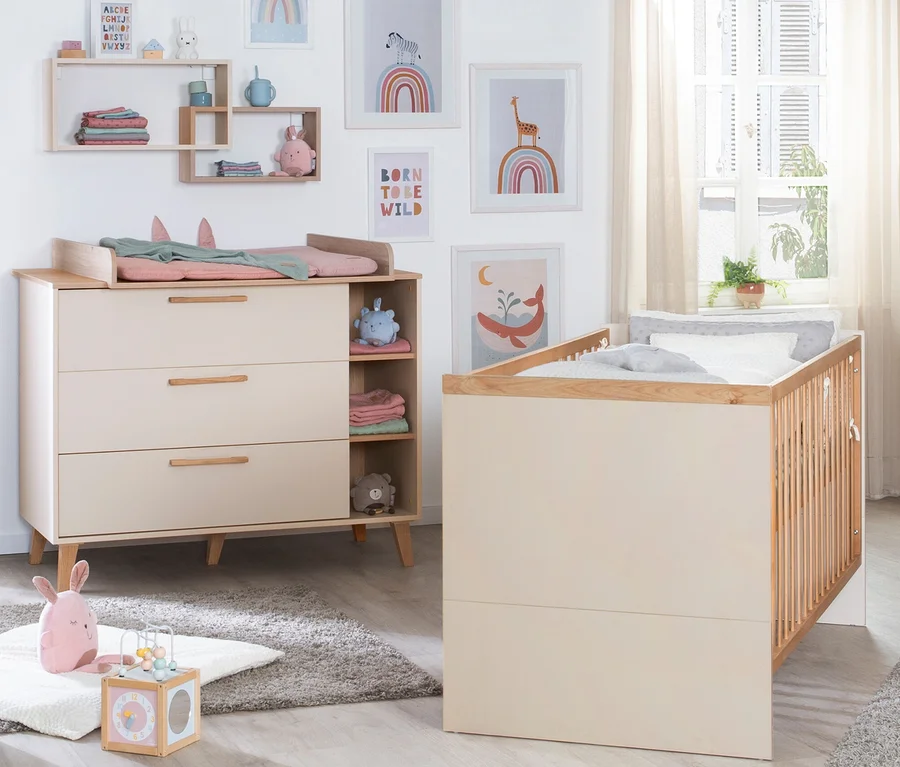 Ein helles Babyzimmer mit einer beigen Wickelkommode, einer rosa Wickelauflage und einem passenden Gitterbett. Wandregale halten Babyartikel, und gerahmte Kunst mit Regenbögen und Tieren schmückt die Wand. Ein rosa Plüschhase und ein hölzerner Aktivitätswürfel liegen auf einem grauen Teppich.