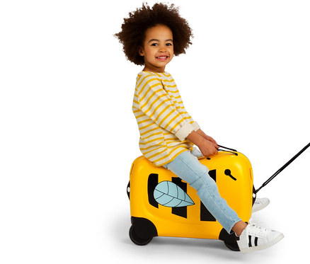 Ein lächelndes Kind mit lockigem Haar sitzt auf einem gelben American Tourister Funrider Aufsitzkoffer im Bienen-Design.