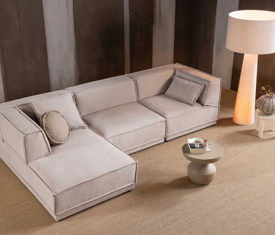 L-förmiges, modulares Sofa in Beige mit Kissen, runder Couchtisch, Stehlampe und Hocker mit Vase auf beigem Teppich.
