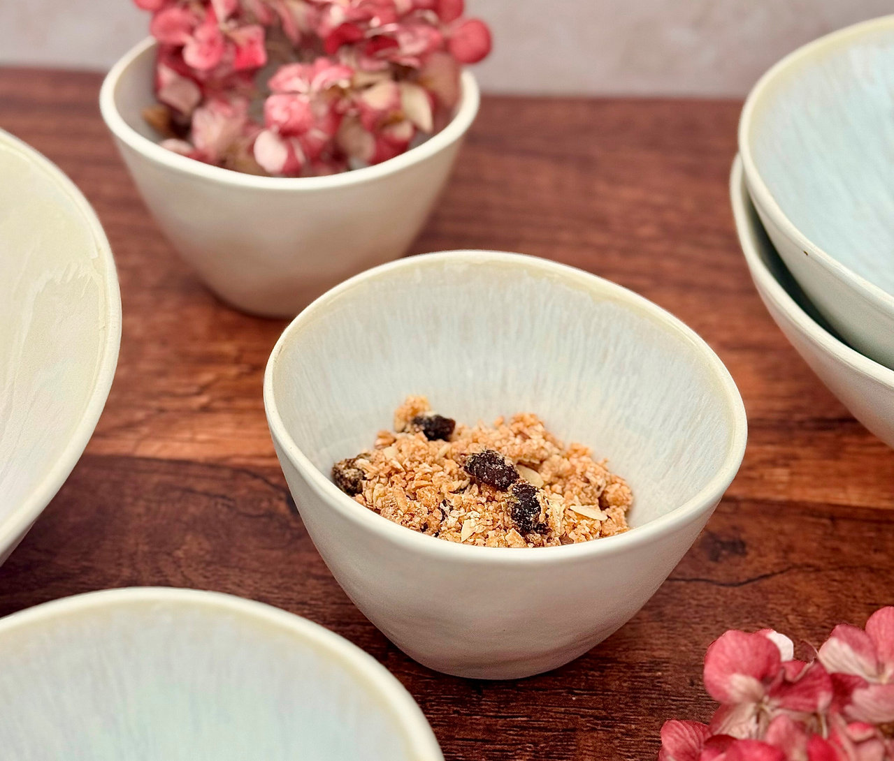 Eine helle Schüssel aus dem Mäser Schüssel-Set »Frozen« mit Müsli und Rosinen auf einem Holztisch, weitere Schüsseln und getrocknete Blumen im Hintergrund.