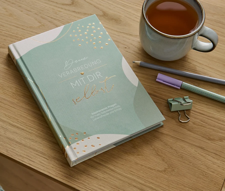 Auf einem Holztisch liegen das Buch »Deine Verabredung mit dir selbst«, eine Tasse Tee, ein Bleistift, ein Textmarker und eine Büroklammer.