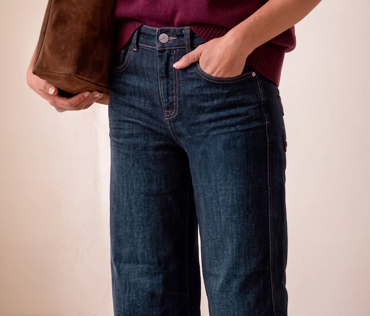 Eine Person trägt dunkelblaue Street One Jeans und einen weinroten Pullover, hält eine braune Wildledertasche und hat eine Hand in der Hosentasche.