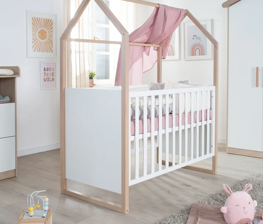 Ein Babyzimmer mit einem weißen Gitterbett mit hellem Holzrahmen in Hausform und rosa Bettwäsche, über dem ein rosa Baldachin drapiert ist. Links steht eine Wickelkommode, rechts ein Kleiderschrank. Auf dem Boden liegt ein grauer Teppich mit einem rosa Hasen und Holzbausteinen. Wandbilder zeigen Sonne und Regenbogen.
