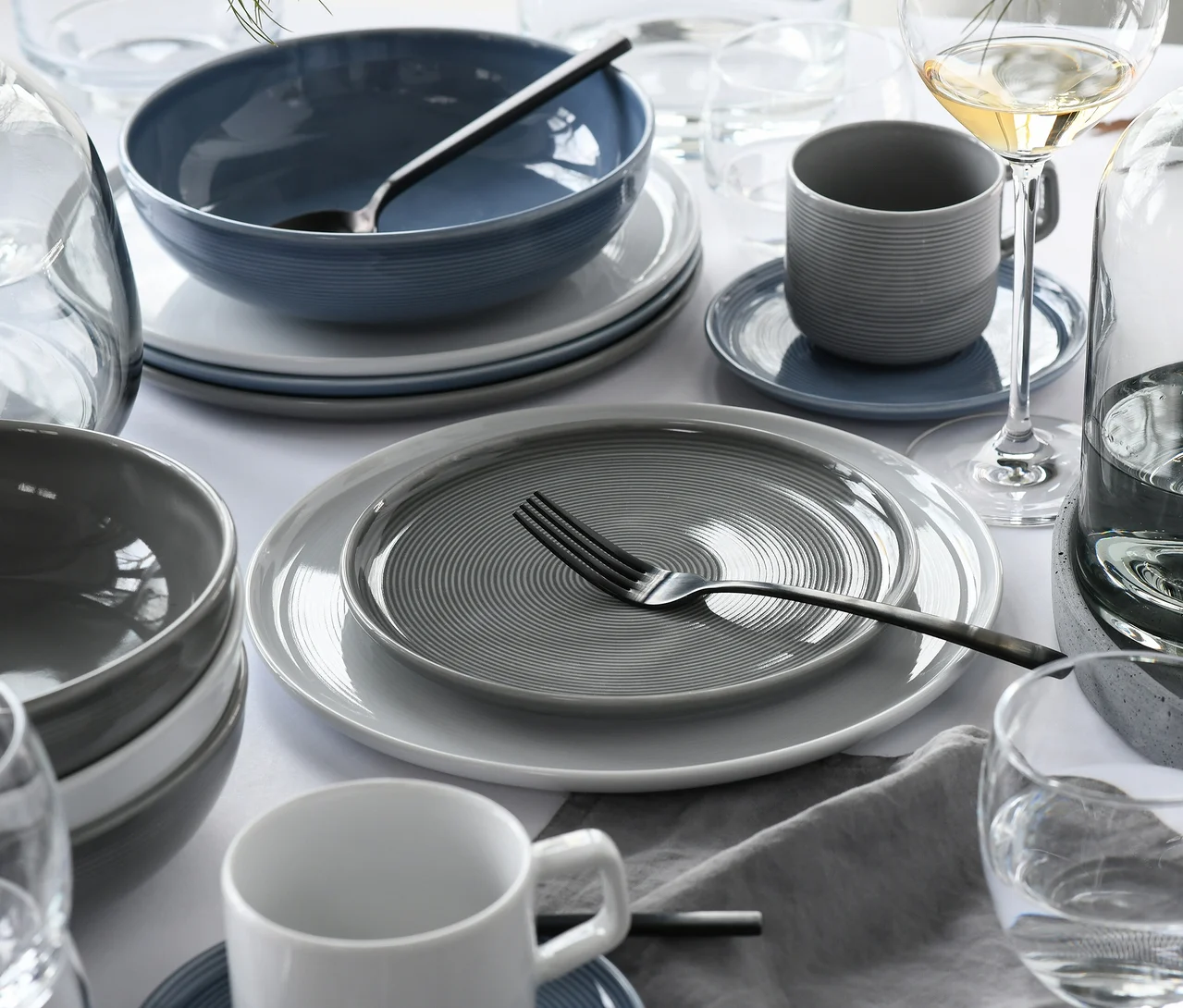 Gedeckter Esstisch mit modernem creaTable Service »Session Urban« Geschirr. Ein grauer Teller mit Spiralmuster und dunkler Gabel liegt auf einem weißen Teller. Stapel von grauen und blauen Schüsseln, eine gerillte graue Tasse auf blauer Untertasse und eine weiße Tasse sind sichtbar. Gläser und ein Weinglas mit Weißwein ergänzen das Arrangement.