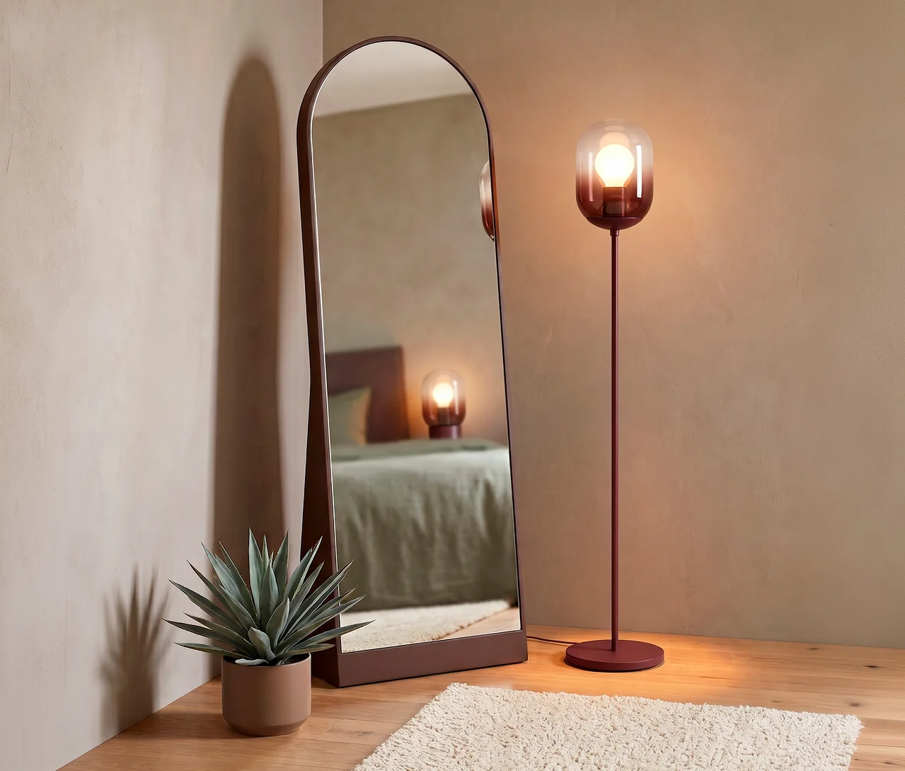 Ein braun gerahmter WOOOD Ganzkörperspiegel »Solari» lehnt an einer hellbraunen Wand und spiegelt ein Bett wider. Rechts davon steht eine rotbraune Stehlampe mit einem Glasfarbverlaufsschirm. Eine Topfpflanze und ein cremefarbener Hochflorteppich liegen auf dem Holzboden.