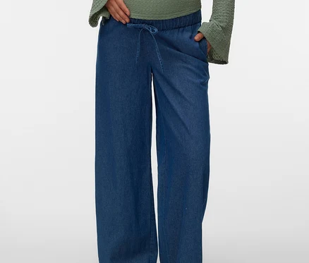 Eine schwangere Person mit der Hand am Bauch trägt eine grüne, langärmelige Bluse und eine blaue MAMALICIOUS weite Jeans »MLJAMAICA« mit Kordelzug, dazu braune Mules.