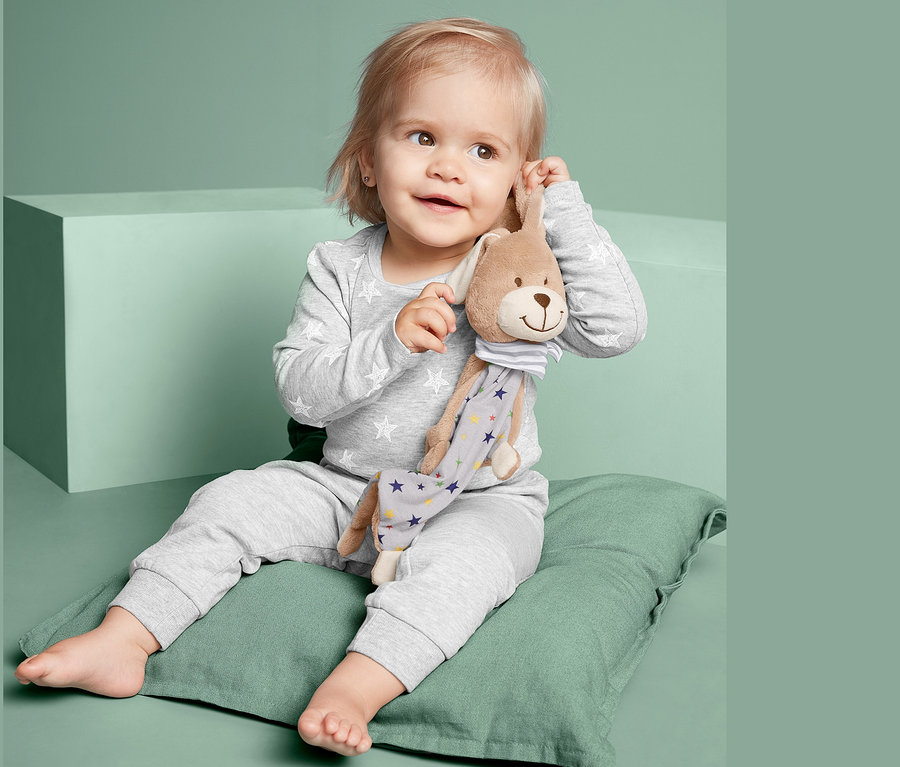 Kleinkind sitzt auf grünem Kissen und hält Schnuffeltuch »Hase«.