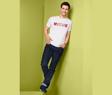 Mann lehnt an einer Wand und trägt eine »Mustang« Jeans in super dark und ein weißes T-Shirt.